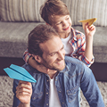 Two people hold paper airplanes, smiling, seated in a cozy living room with a couch in the background.
